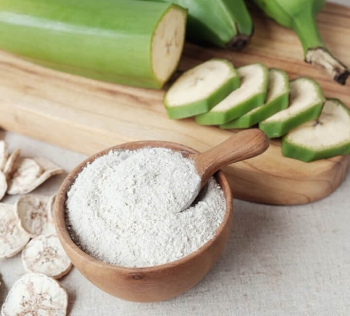Green banana powder in a wooden bowl with sliced green bananas